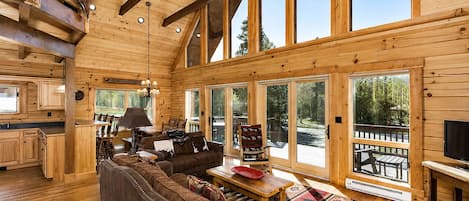 A log cabin interior with a large wooden ceiling, a cozy living area with a sofa and chairs, a dining table, and a TV.