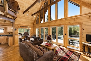 Ein Blockhaus-Interieur mit einer großen Holzbalkendecke, einer gemütlichen Wohnbereits mit Sofa und Sesseln, einem Esstisch und einem Fernseher.