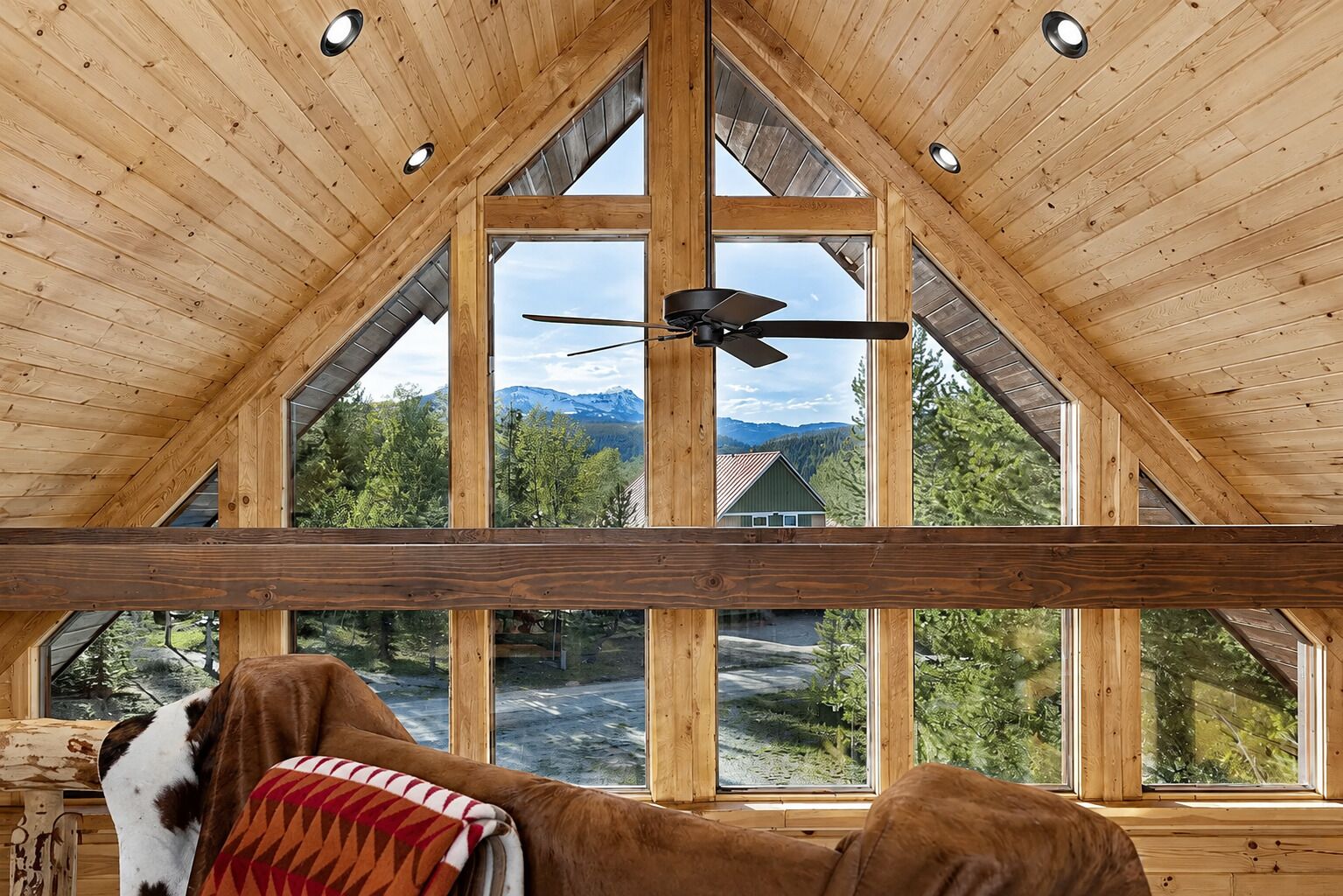 Un intérieur en bois, de grandes fenêtres, un ventilateur de plafond et une vue sur un paysage montagneux.