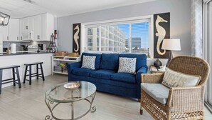 Un salon moderne avec un canapé bleu, une table basse en verre, un fauteuil en osier et une cuisine en arrière-plan.