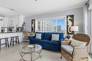 Ein modernes Wohnzimmer mit einem blauen Sofa, einem Glastisch, einem Korbstuhl und einer Küche im Hintergrund.