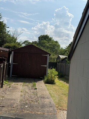 A wooden shed with a metal door, a concrete pathway leading to it, and a grassy yard with a fence in the background.