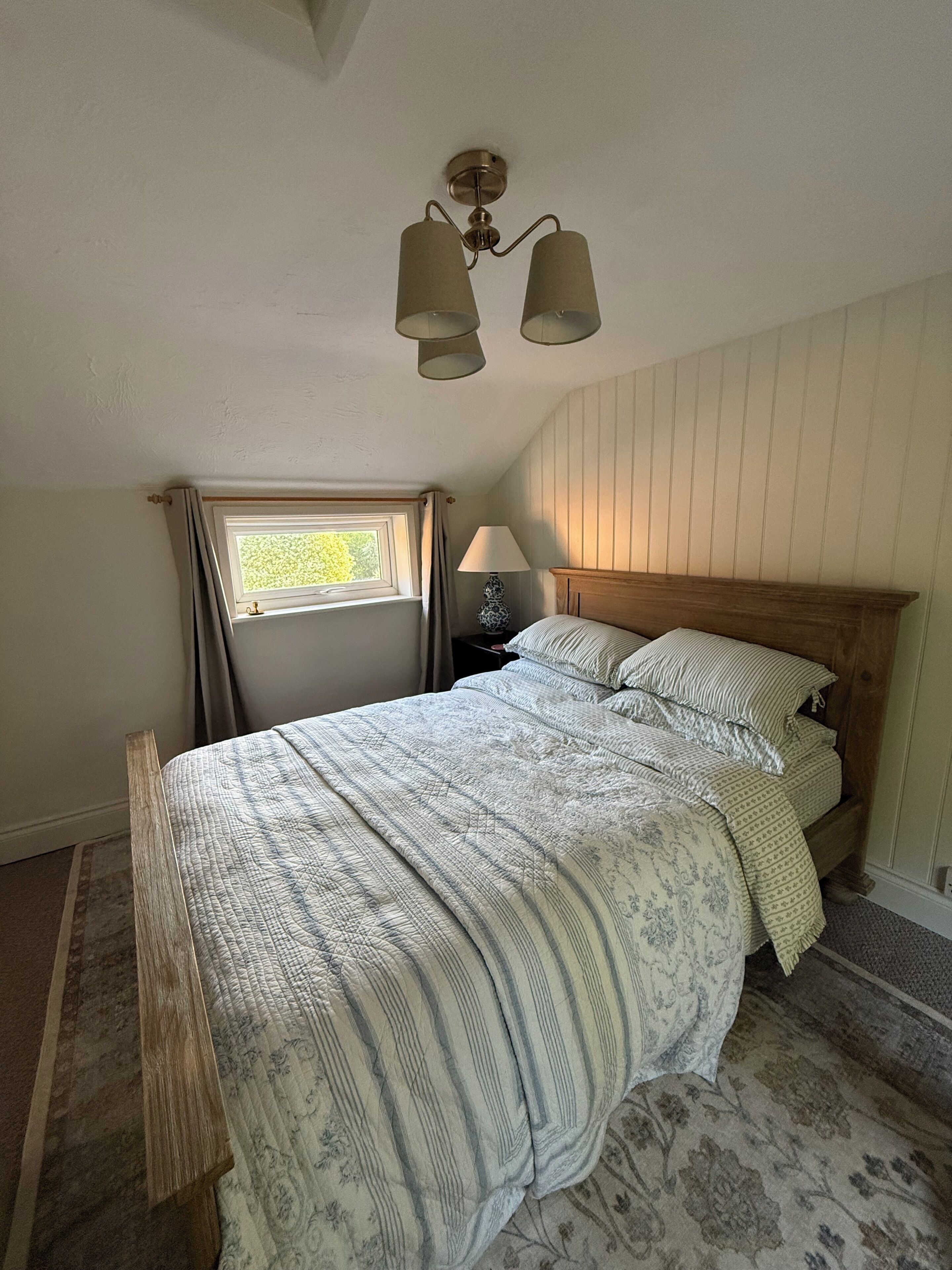 A bedroom with a wooden bed, a nightstand, a lamp, a window with curtains, and a ceiling fan.