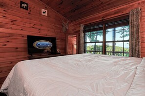 A wooden cabin bedroom with a bed, a mounted television, and a window with a view of trees. - A View For Two | Ellijay, GA (Gilmer County)