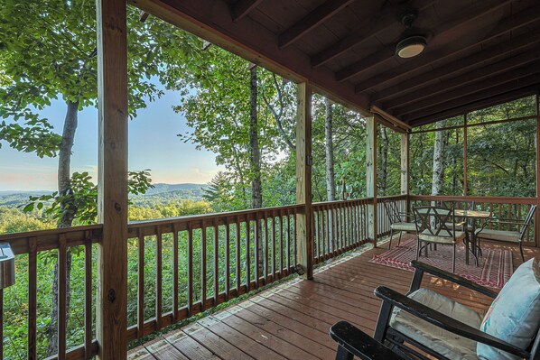 A wooden deck with a view of a forest and mountains, a table with chairs, and a hanging light fixture. - A View For Two | Ellijay, GA (Gilmer County)