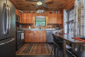 A kitchen with wooden cabinets, stainless steel appliances, a window with blinds, and a ceiling fan. - A View For Two | Ellijay, GA (Gilmer County)