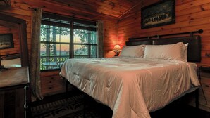 Quarto de cabana de madeira com uma cama, uma mesa de cabeceira, um abajur, uma janela com cortinas e um quadro na parede.
