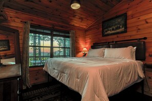 Ein Schlafzimmer einer Holzhütte mit einem Bett, einem Nachttisch, einer Lampe, einem Fenster mit Vorhängen und einem gerahmten Bild an der Wand.