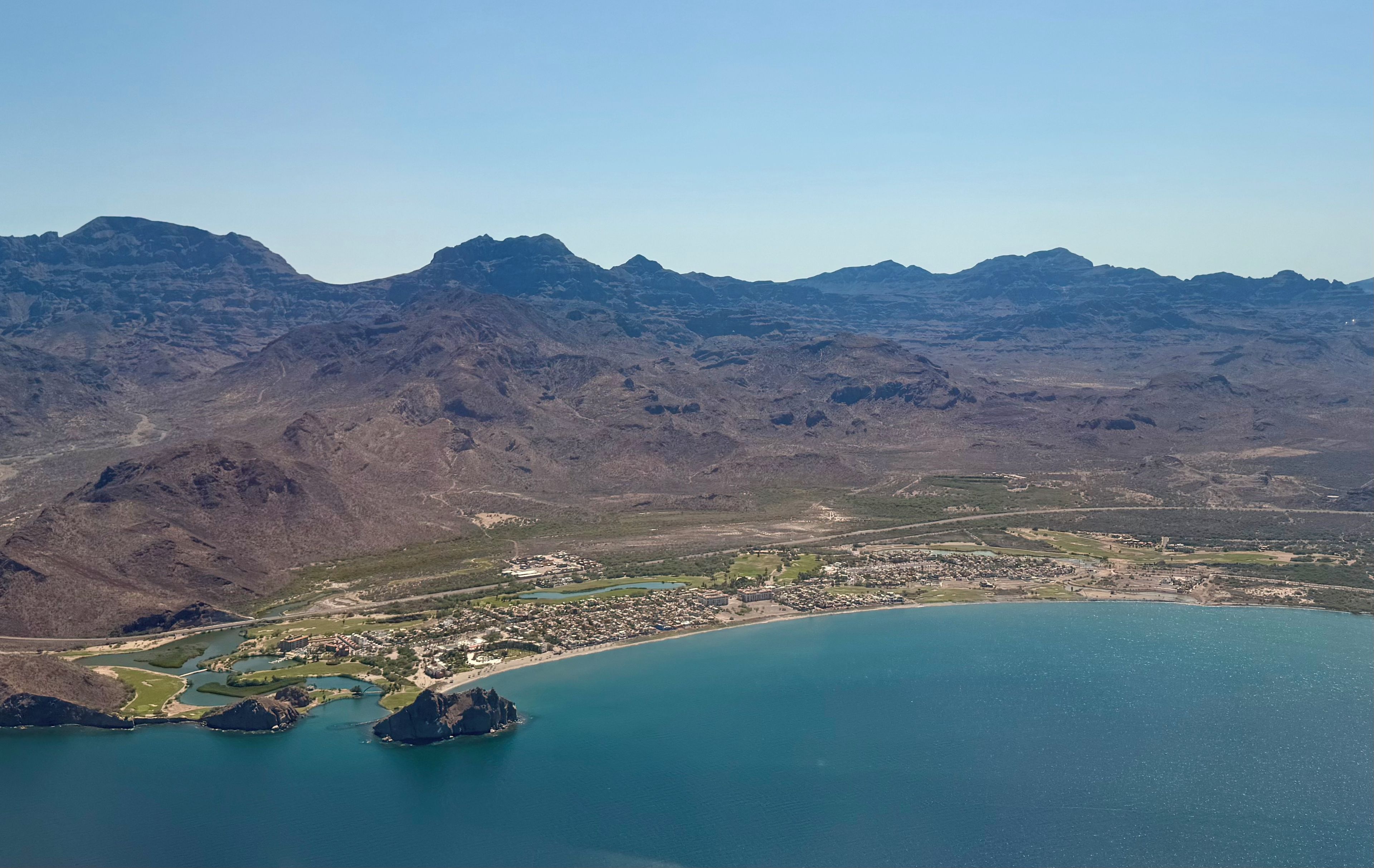 An aerial view of a coastal town with mountains in the background, a marina, and a bridge connecting the town to the mainland.