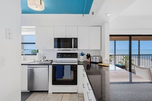 Eine moderne Küche mit weißen Schränken, einem Backofen, einer Mikrowelle und einem Toaster. Große Fenster bieten Blick auf den Ozean.