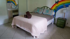 Une chambre à coucher avec un lit, des oreillers, une couverture, une table de chevet verte, une lampe et un mur décoré d’un arc-en-ciel et de motifs d’animaux.