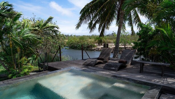 Une piscine entourée de végétation tropicale et bordée d’une étendue d’eau, avec des chaises longues et une terrasse en bois.
