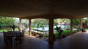Une terrasse extérieure couverte, dotée d’une table et de chaises, d’un hamac, et offrant une vue sur une piscine et sur la verdure.