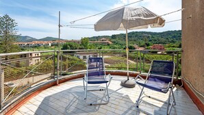 A balcony with two blue chairs, a white umbrella, and a view of buildings and hills.
