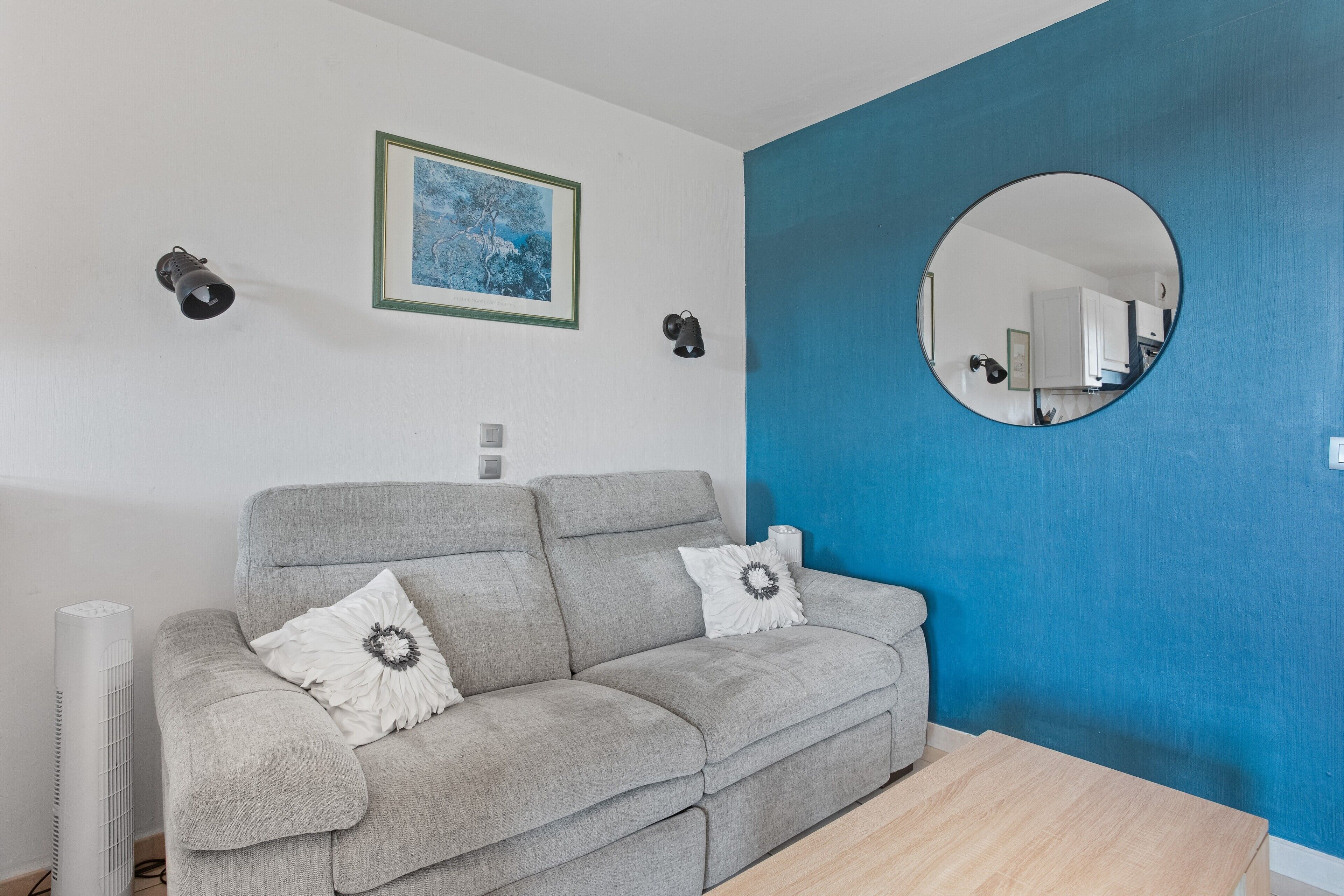 A modern living room with a grey sofa, a round mirror, a framed picture, and a standing heater.