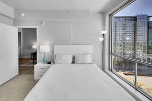 A modern bedroom with a large bed, a nightstand, a lamp, and a view of a cityscape through a large window. - Your World Cup Stay! Furnished 1BR nr Levi Stadium (San Jose)