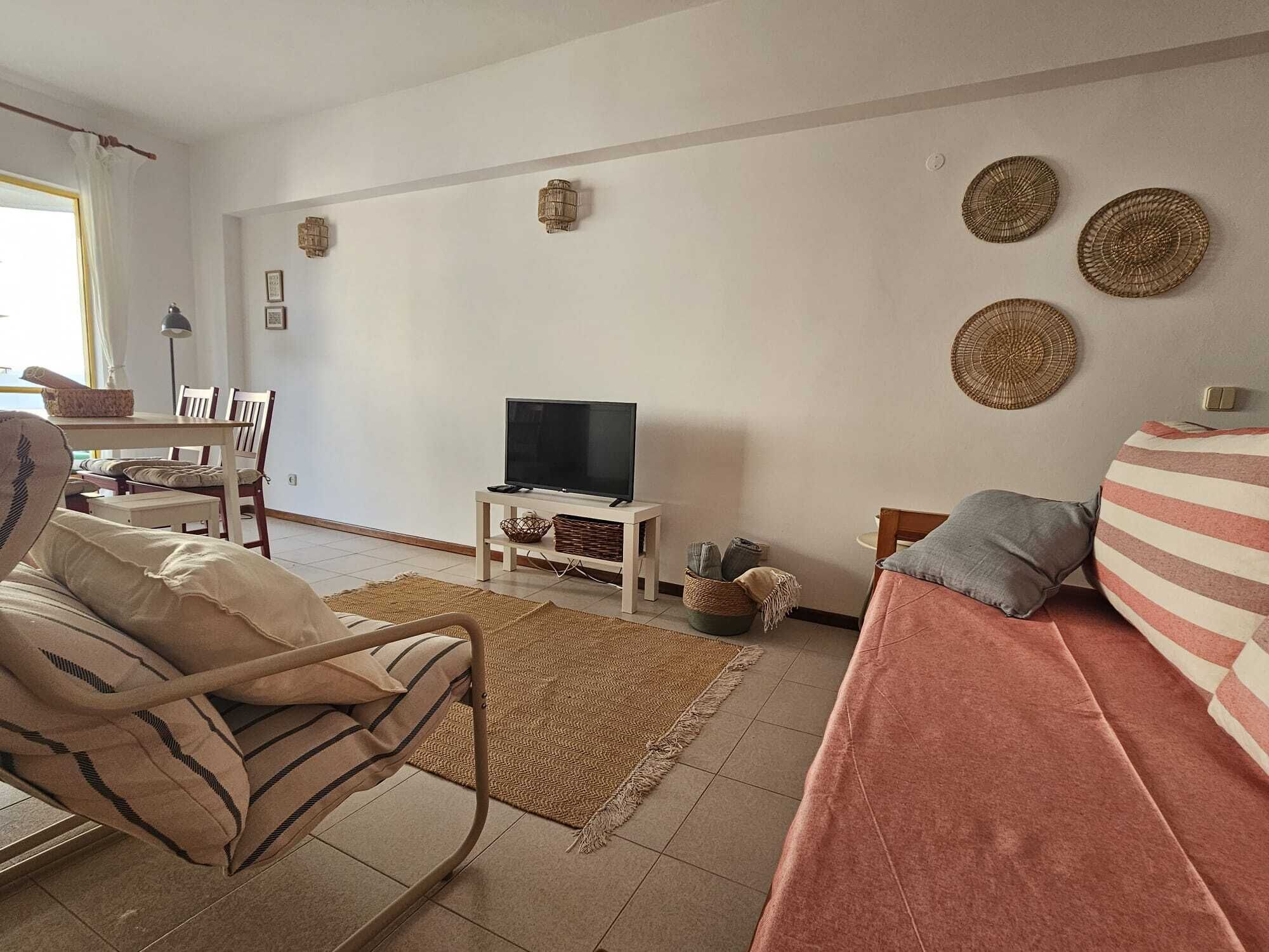 Ein Wohnzimmer mit Sofa, Sessel, Fernseher auf einem Schrank, Teppich und Wandbildern.