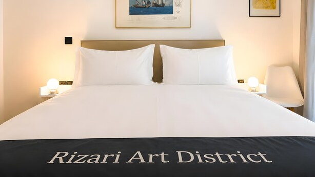 Ein ordentlich bezogenes Bett mit weißen Leinen, ein gerahmtes Kunstwerk darüber und eine Lampe auf jeder Seite.