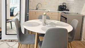 Eine moderne Küche mit Kochinsel, ein Essbereich mit rundem Tisch und Stühlen, und Blick in ein Schlafzimmer durch eine geöffnete Tür.