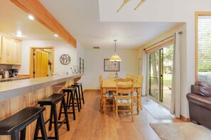 Una cocina con armarios de madera, una zona con taburetes de bar, una mesa de comedor con sillas y un amplio ventanal corredero que da a un área exterior.
