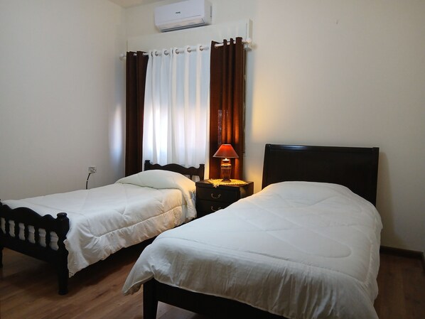 A room with two single beds, a bedside table with a lamp, a power outlet, and an air conditioning unit on the wall.