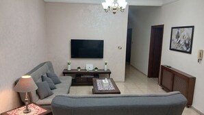 Uma sala de estar com um sofá cinzento, uma mesa de centro, uma televisão na parede, um candelabro e uma obra de arte emoldurada na parede.