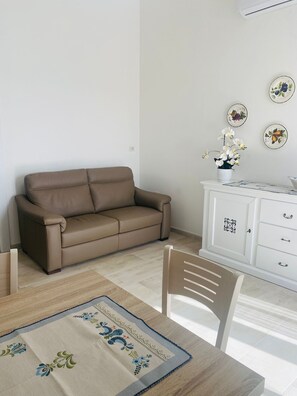 Una stanza con un divano marrone, un armadietto bianco, un tavolo da pranzo con tovaglia floreale e piatti decorativi appesi al muro.