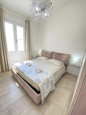 A bedroom with a bed, curtains, a chandelier, a bedside table with a lamp, and a window.