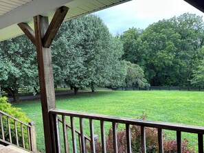 Eine Veranda mit Holzgeländer, die auf eine grüne Fläche mit Bäumen und einem Zaun blickt.