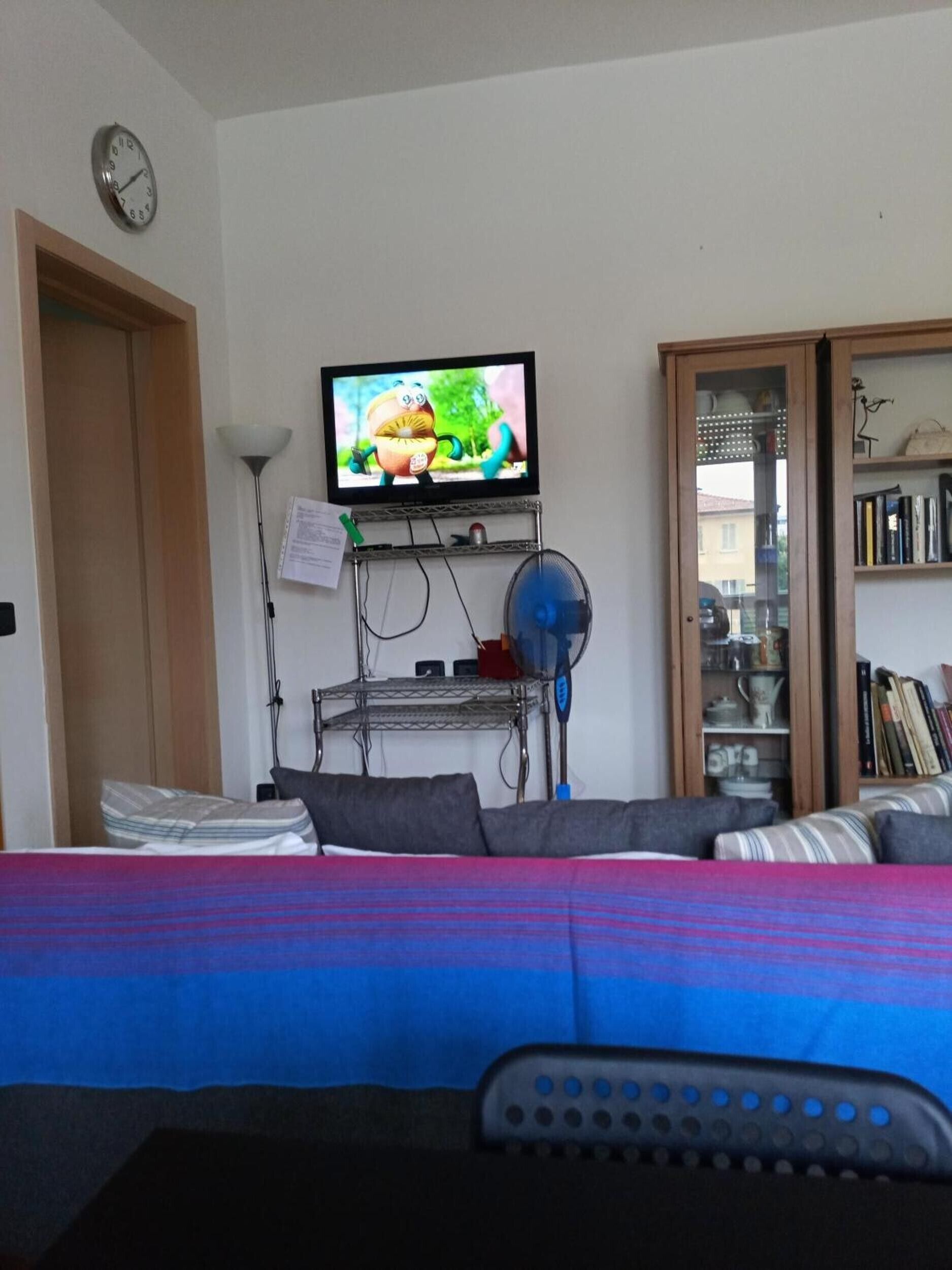 A living room with a television mounted on the wall, a bookshelf, a clock, and a bed with a colorful blanket.