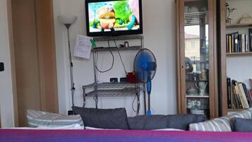 A living room with a television mounted on the wall, a bookshelf, a clock, and a bed with a colorful blanket.