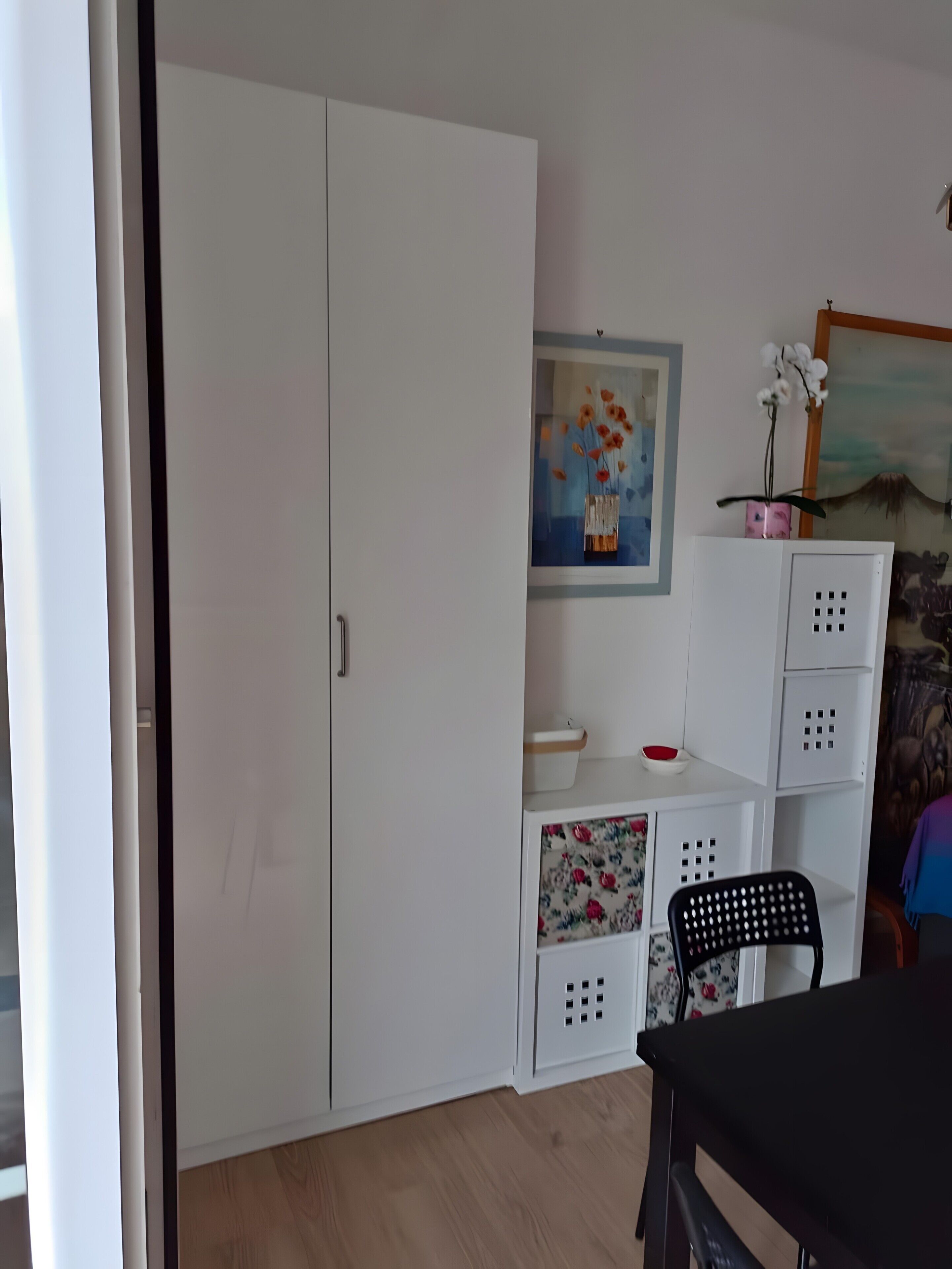 A room with a white wardrobe, a painting of flowers, a small table with a chair, and a decorative plant.