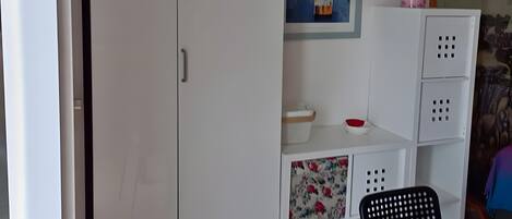 A room with a white wardrobe, a painting of flowers, a small table with a chair, and a decorative plant.
