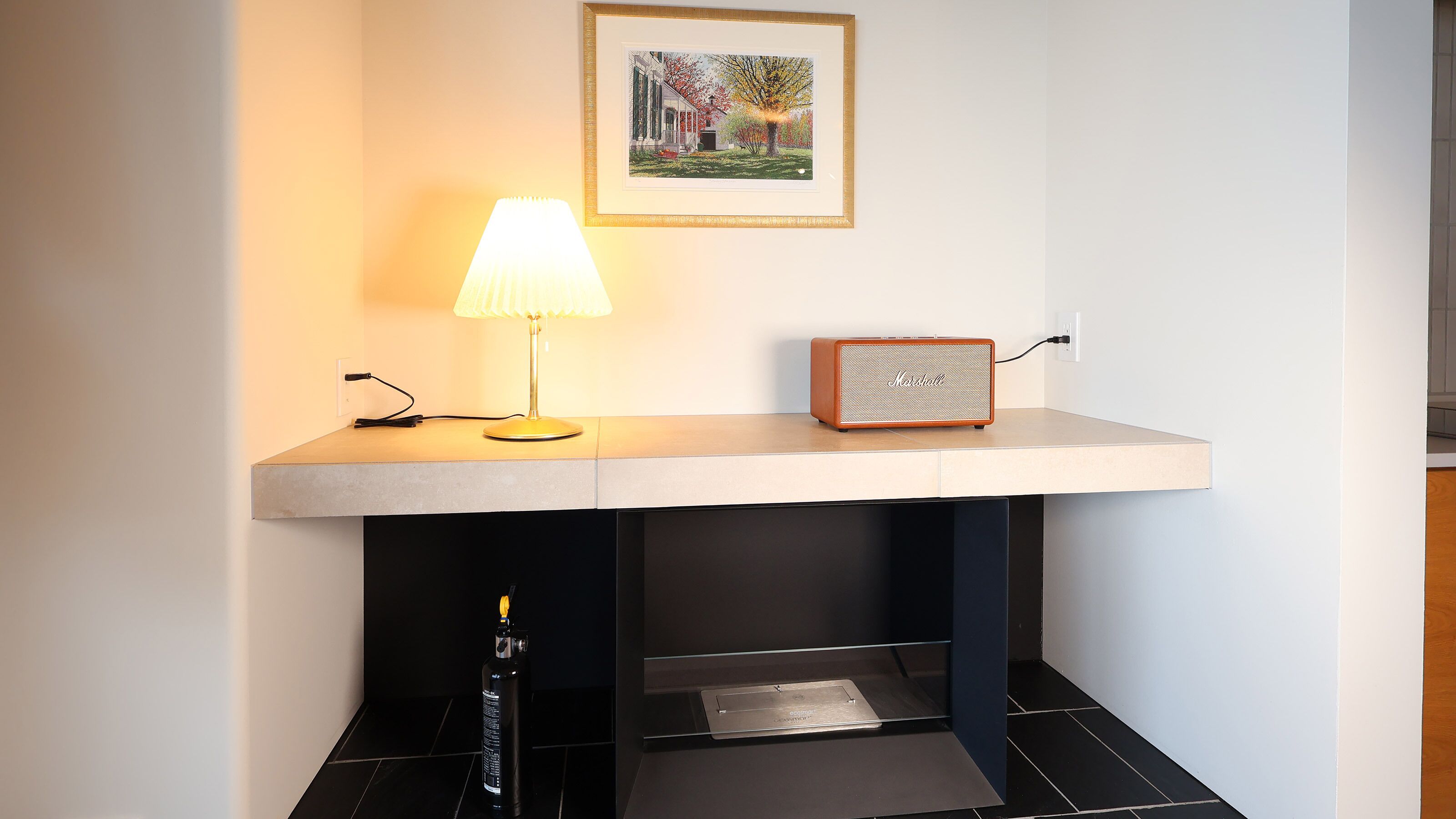 A room with a desk, a lamp, a speaker, and a framed picture on the wall.