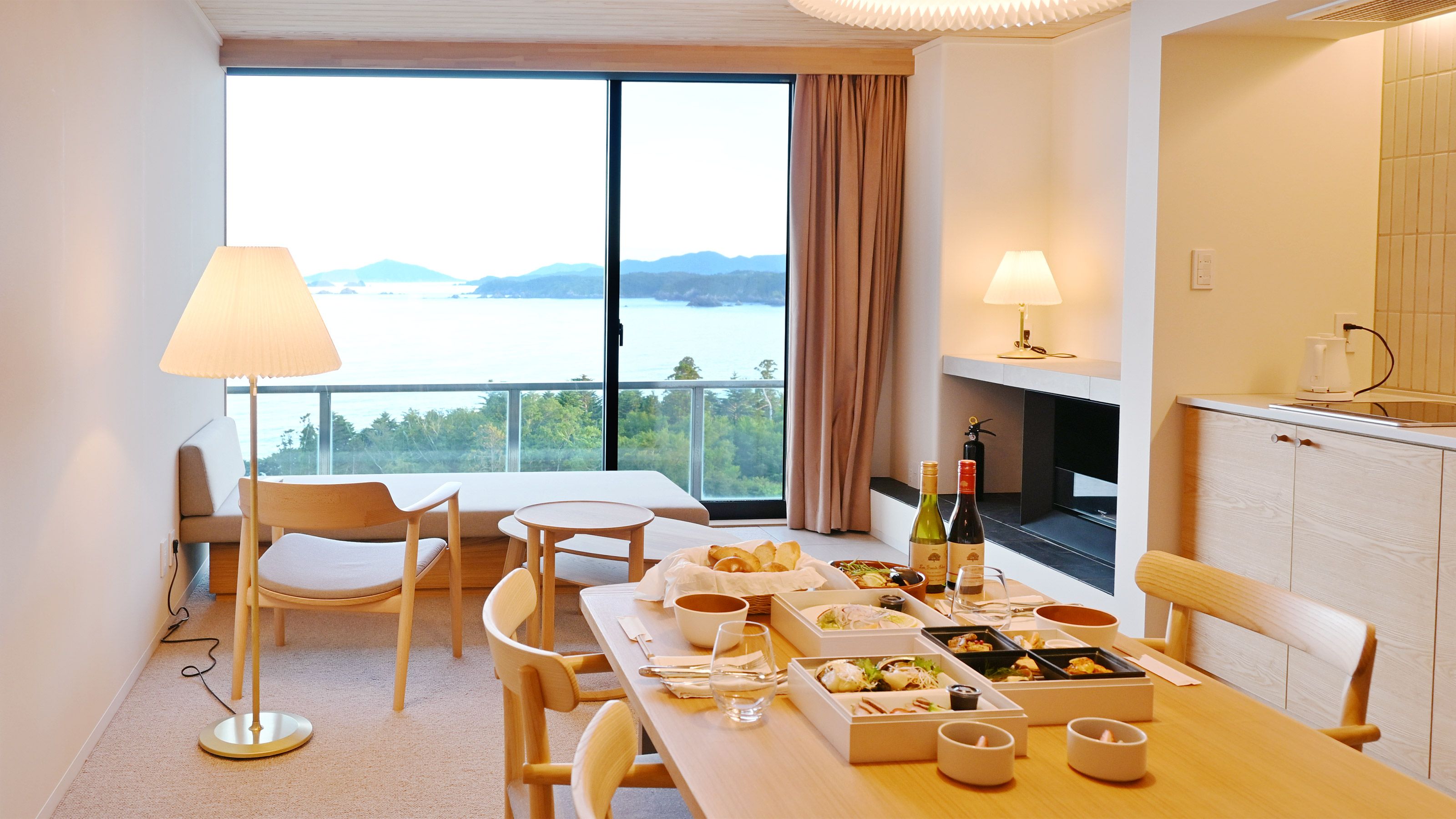 A hotel room with a dining table set for two, a sofa, a view of the sea, and a fireplace.