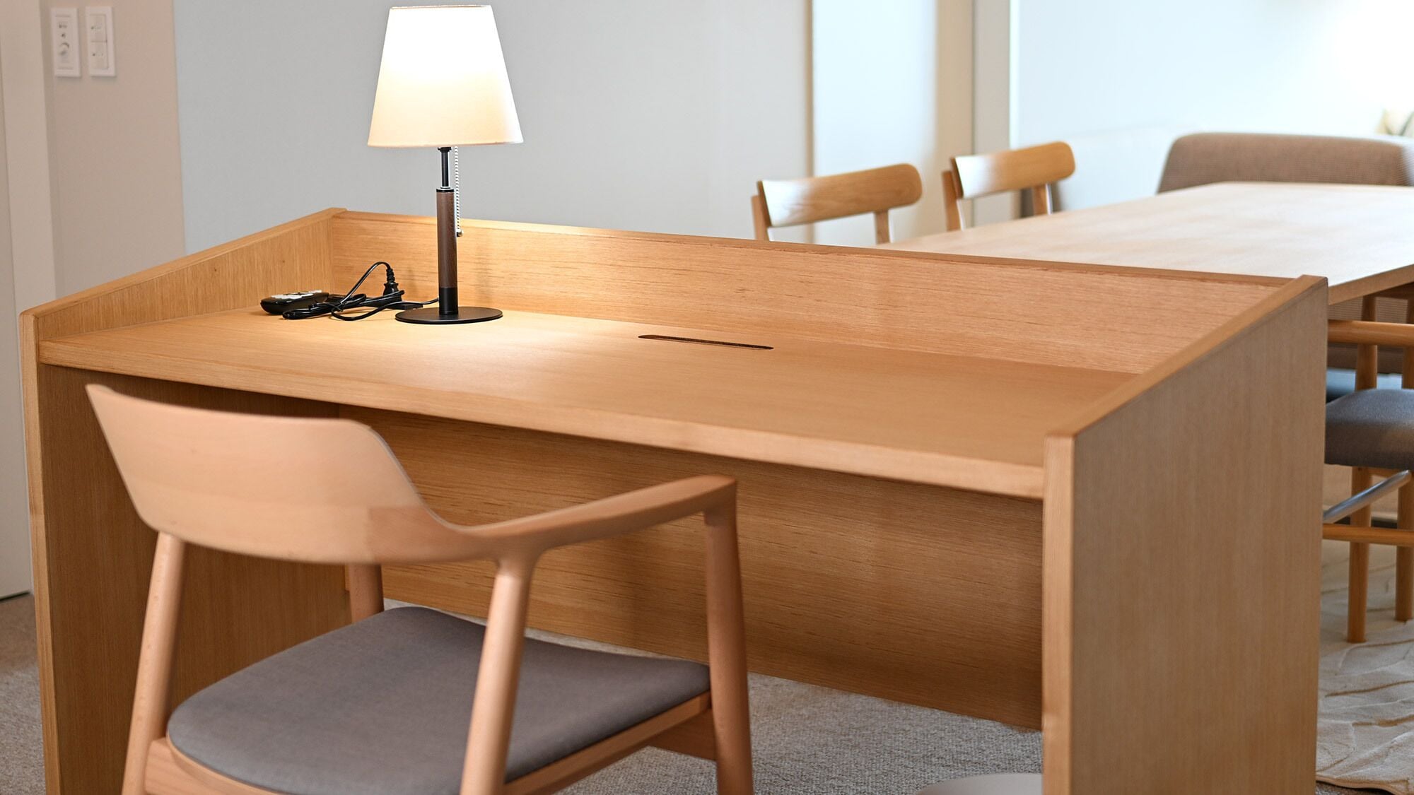 A wooden desk with a lamp, a chair, and a table in a room with a carpeted floor.