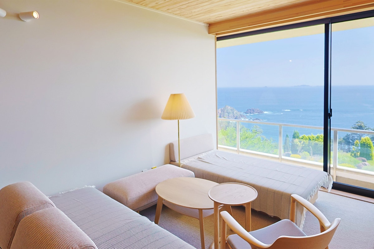 Quarto de hotel com cama, uma pequena mesa, uma cadeira, abajur e vista para o mar.