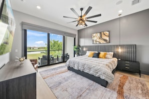 A modern bedroom with a bed, a ceiling fan, a nightstand, a desk, and a large window with a view of greenery. - WOW Home with Arcade Floor + Theater | Private Pool & Spa (Kissimmee)