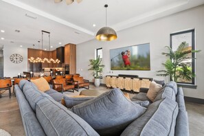 A modern living room with a large gray sofa, wooden dining table, and a TV mounted on the wall. - WOW Home with Arcade Floor + Theater | Private Pool & Spa (Kissimmee)