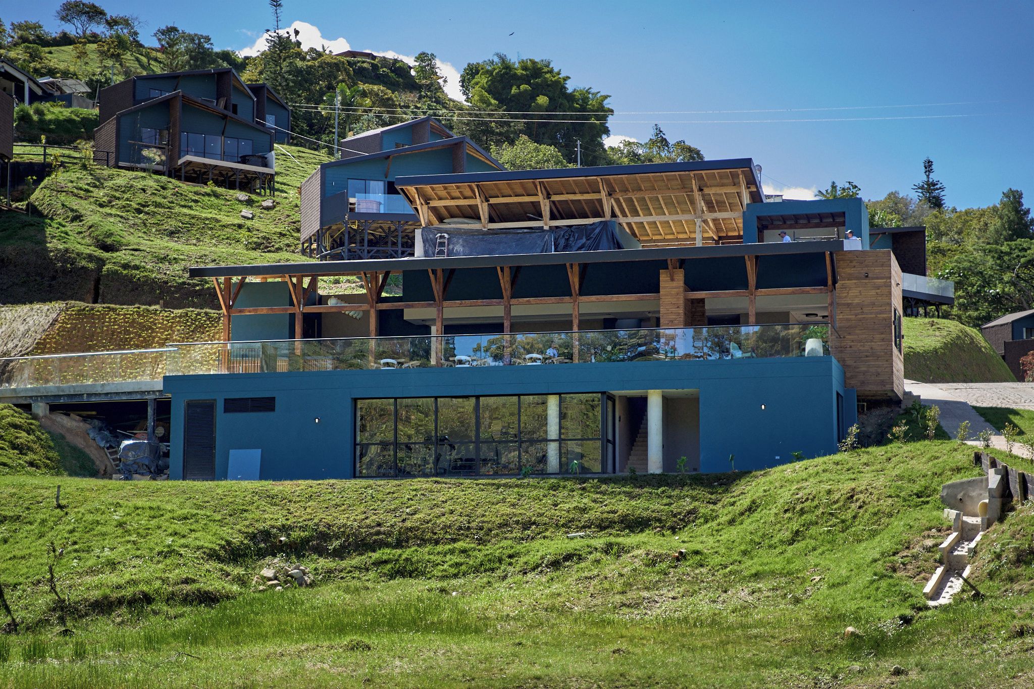 A modern multi-story building with a flat roof and large windows, surrounded by greenery and hills.