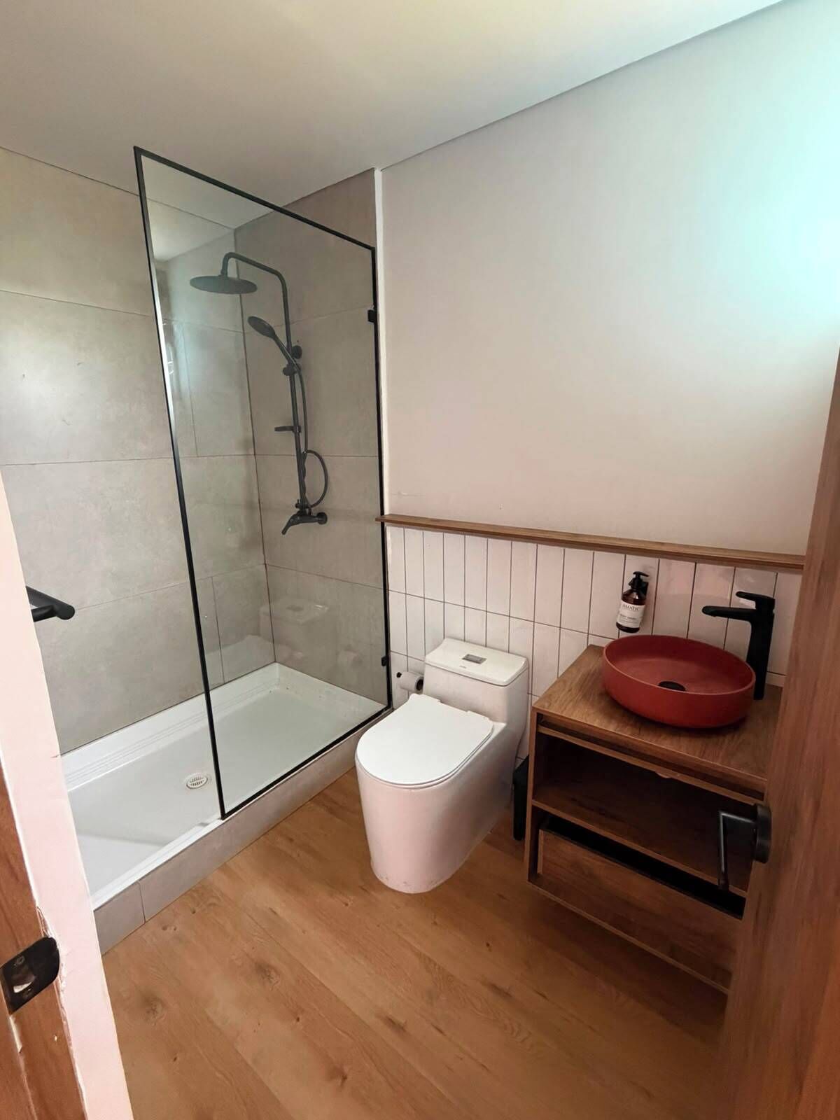 A bathroom with a glass-enclosed shower, a toilet, and a wooden vanity with a red sink.