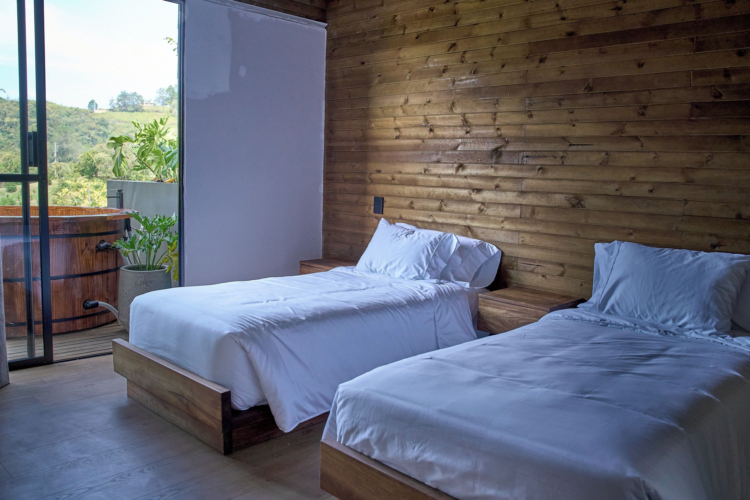 A bedroom with two beds, wooden walls, and a view of greenery outside.