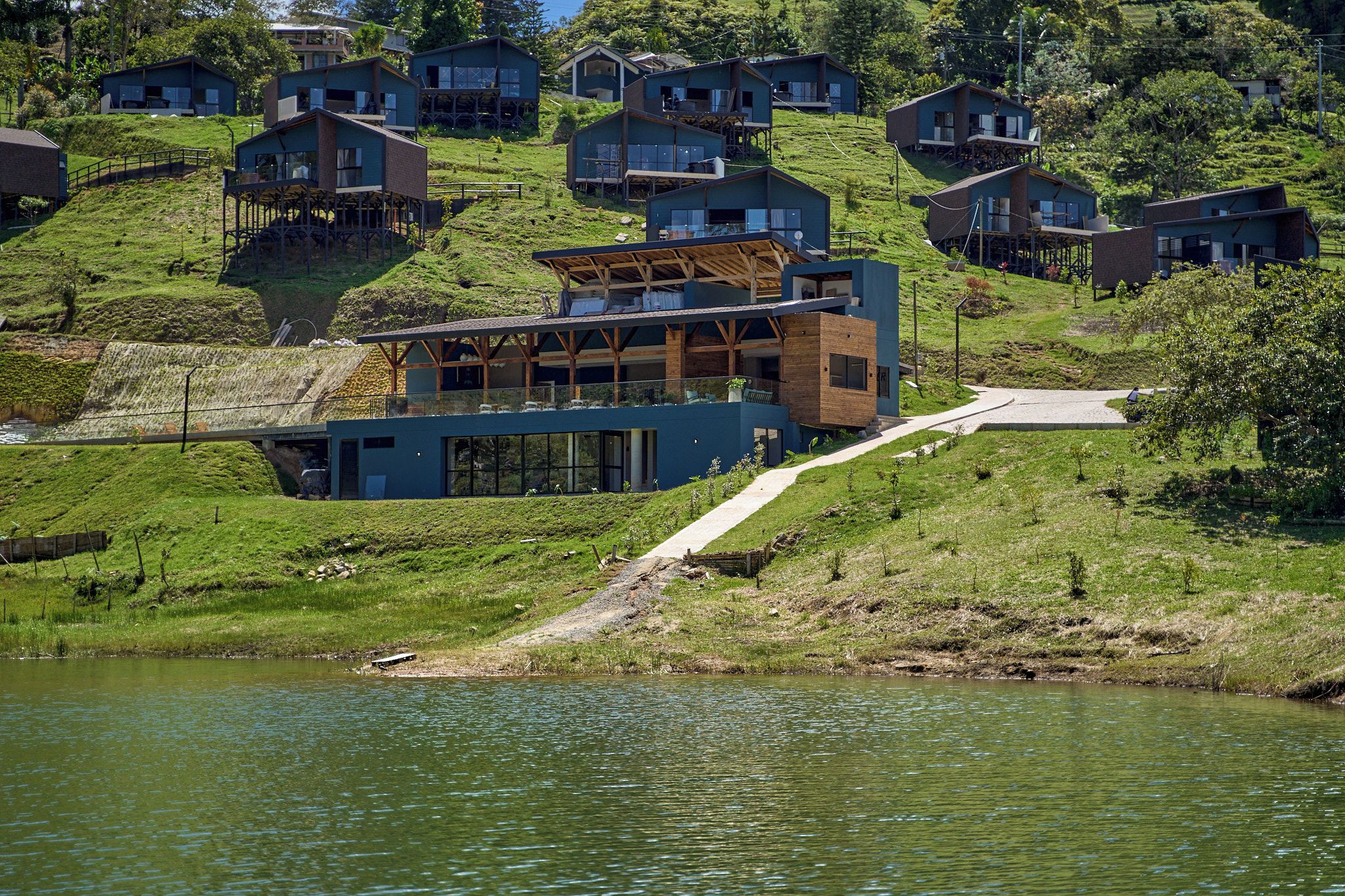 Un edificio moderno con un amplio frente de vidrio, ubicado en una colina con vista a un cuerpo de agua.