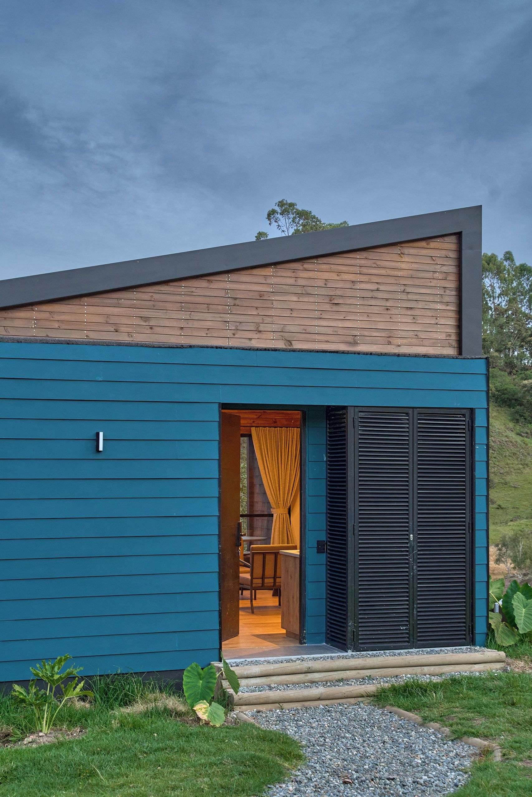 Una casa moderna con fachada azul, techo inclinado de madera y una puerta con contraventanas negras.