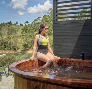 Una persona sentada en un jacuzzi al aire libre, con una terraza de madera y vistas a un paisaje verde.
