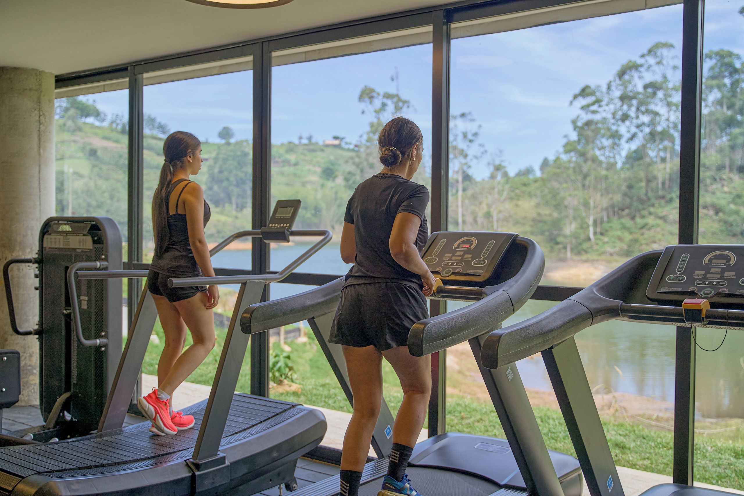 Duas pessoas a correr em esteiras com vista para uma paisagem natural através de grandes janelas.