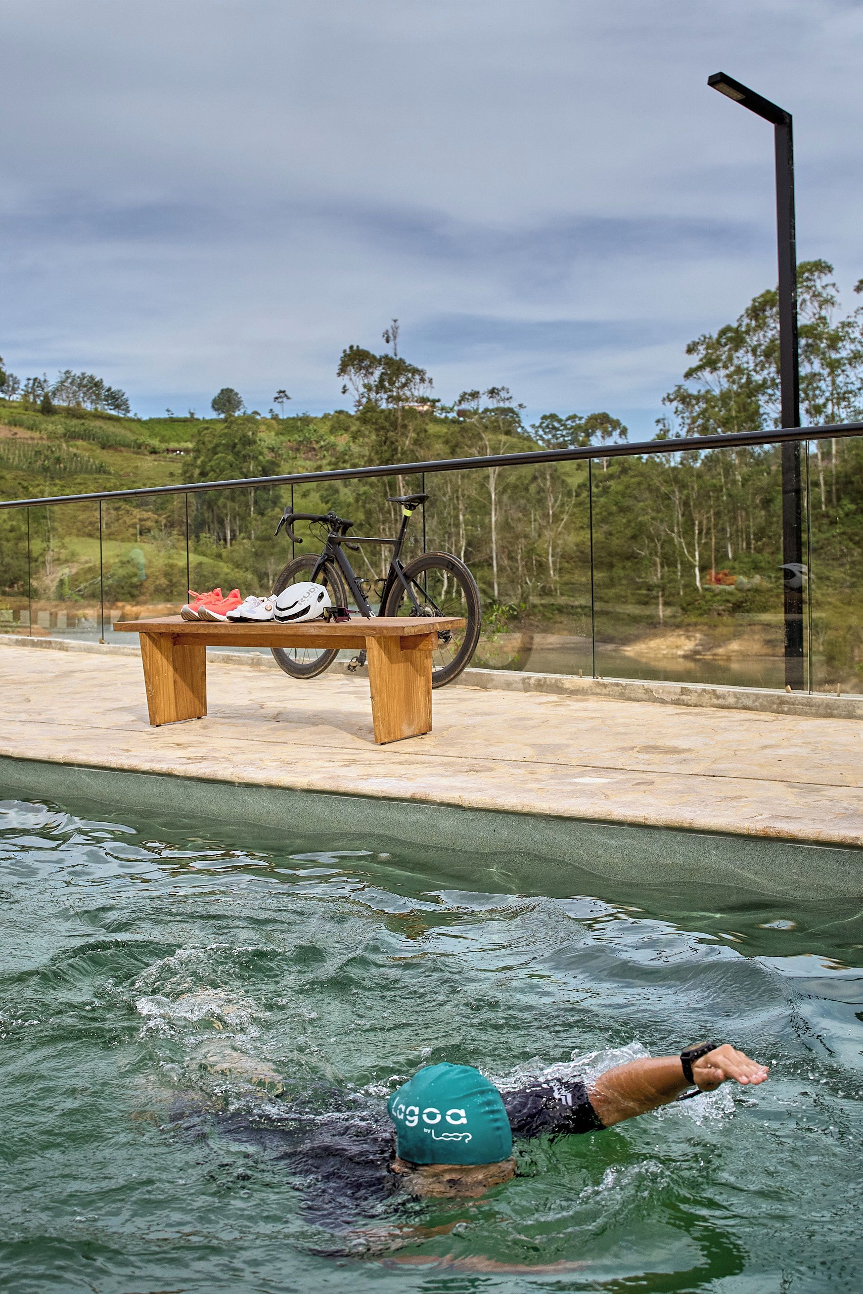 A swimming pool with a person in the water, a bicycle, and a bench with shoes and a helmet.