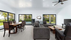 A modern living room with a dining area, large windows, a ceiling fan, and a TV.