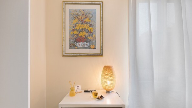 Et værelse med et hvidt bord, en gul stol, et indrammet kunstværk, en lampe og en lille vase med et lys.