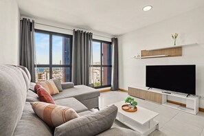 Un salon moderne avec un canapé gris, un téléviseur à écran plat et une vue sur la ville grâce à de grandes fenêtres.